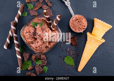 Schaufeln Sie Schokoladeneis in einer Glasschüssel mit Waffelstäbchen, Kegel und Schokolade auf einem schwarzen Schiefer Stockfoto