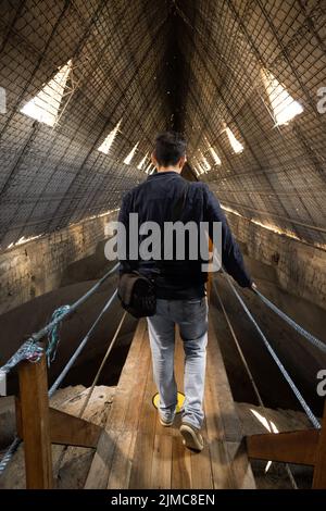 Person von hinten auf einer Holzbrücke zu Fuß, trägt eine Jacke und Jeans, hat einen Koffer, Interieur mit Industriearchitektur, touristischen Lebensstil Stockfoto