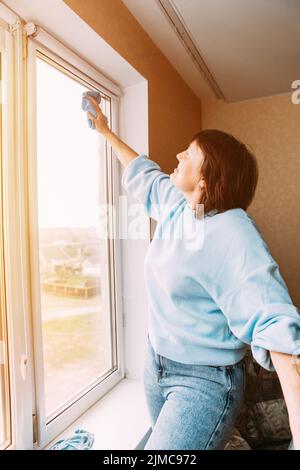 Glücklich Schüchtern Positive Reife Frau Reinigung Fenster. Konzept Für Aktive Senioren. Ältere Frau Reinigung Apartment. Hausfrau. Kaukasische Frau Im Mittleren Alter Stockfoto