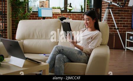Glückliche asiatische Frau mit modernen Tablet im Internet surfen, während auf der Couch zu Hause sitzen. Fröhlicher, herzlicher Mensch mit Touchscreen-Gerät, der Webseiten surft, während er Remote arbeitet. Stockfoto