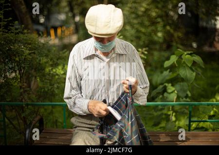 Der alte Mann sitzt im Sommer auf der Bank. Rentner in Russland in der Nähe des Hauses. Mann mit medizinischer Maske. Opa in Hemd und Mütze. Stockfoto