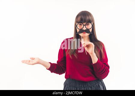 Frau mit langen, geraden braunen Haaren, die ihr Gesicht mit einem gefälschten Schnurrbart aus Papier und einer Brille auf einem Stock bedeckt, lächelt und schaut auf die Kamera auf weißem Hintergrund. Hochwertige Fotos Stockfoto