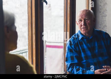 Kaukasisches europäisches Seniorenpaar zu Hause sitzt im Winter in der Nähe des frostigen Fensters und Ehemann spricht mit seiner Frau. Hochwertige Fotos Stockfoto