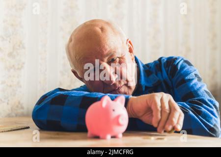 Trauriger, verlorener und deprimierter kaukasischer älterer Mann, der mit einem Sparschwein auf dem Tisch lag und seine letzten Münzen zählte. Hochwertige Fotos Stockfoto
