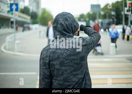 Unbekannte Person schießt auf dem Smartphone auf der Straße. Person beobachtet jemanden. Kerl jagt Mädchen. Seltsamer Passant in der Stadt. Student in Hood. Stockfoto