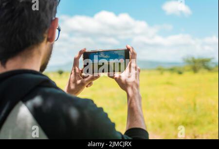 Rückansicht einer Person, die mit seinem Handy ein Foto eines Feldes macht, Rückansicht eines Mannes, der Fotos eines Feldes macht, Nahaufnahme eines Touristen-Mannes, der fotografiert Stockfoto