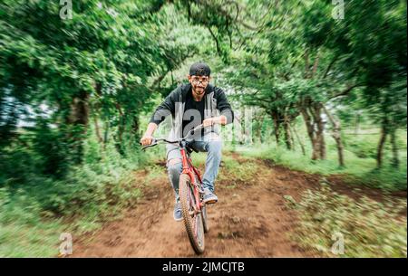 Porträt eines Mannes in Cap, der auf einer Landstraße ein Fahrrad fährt, einer Person, die auf dem Land ein Fahrrad fährt, einer Fahrradfahrerin auf einem Landweg Stockfoto