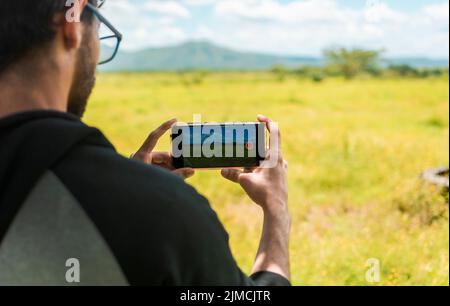Nahaufnahme von Menschenhänden, die mit dem Handy Fotos von einer Landschaft machen. Rückansicht einer Person, die mit seinem Handy ein Foto eines Feldes macht, hinten Stockfoto
