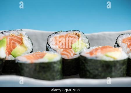 Nahaufnahme von leckeren norwegischen Futomaki-Sushi-Rollen mit Lachs und Avocado, serviert auf blauem Hintergrund im hellen Studio Stockfoto