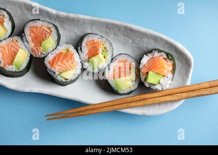 Von oben Set von leckeren Futomaki norwegischen Sushi-Rollen mit Lachs und Avocado serviert in der Nähe von Essstäbchen auf blauem Hintergrund in hellen Studio Stockfoto