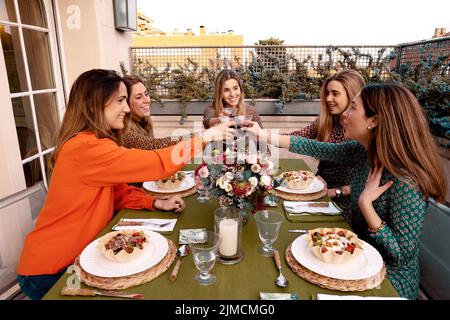 Gruppe positiver Freundinnen, die sich anguckend anguckend und mit Weinläsern toasten, während sie am Tisch mit Salaten auf der Terrasse zu Abend essen Stockfoto