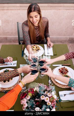 Gruppe positiver Freundinnen, die mit Weinbässen toasten, während sie am Tisch mit Salaten auf der Terrasse essen Stockfoto