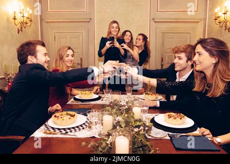 Gruppe positiver Freunde, die sich gegenseitig anguckten und beim Abendessen im Restaurant mit Weingläser am Tisch mit leckerem Essen toasten Stockfoto