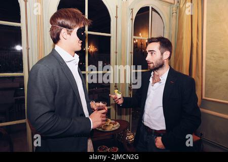 Männliche Freunde in eleganter Kleidung und Maskerade plaudern beim gemeinsamen Abendessen und Weintrinken im Restaurant Stockfoto