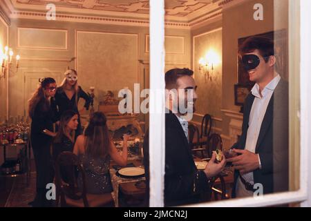 Durch das Fenster einer Gruppe von Freunden in eleganter Kleidung und Maskerade plaudern Masken beim gemeinsamen Abendessen im Restaurant Stockfoto
