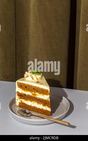 Scheibe von leckeren süßen Karotten Biskuit mit Creme mit Minzblatt verziert serviert auf dem Teller mit Löffel auf dem Tisch in hellen Raum mit Vorhang auf der Rückseite Stockfoto