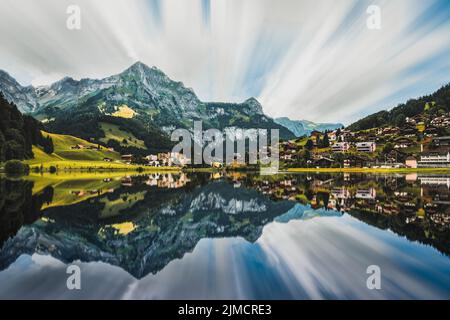 Spektakuläre Landschaft von kleinen Wohnhäusern auf einer Wiese, umgeben von massiven grünen felsigen Bergen, die sich in einem ruhigen See spiegeln Stockfoto