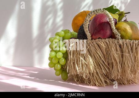 Stillleben von verschiedenen reifen Früchten und Trauben in Korbkorb mit grünen Minzblättern auf dem Tisch vor weißem Hintergrund mit Schatten gelegt Stockfoto