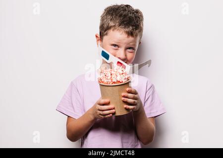 Fröhlicher Junge mit modernen 3D Gläsern, die Pappeimer mit Popcorn halten und die Kamera im Studio vor weißem Hintergrund betrachten