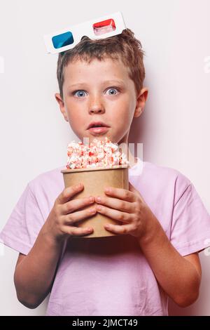 Staunender Junge mit modernen 3D Gläsern, die Pappeimer mit Popcorn halten und die Kamera vor weißem Hintergrund im Studio betrachten Stockfoto