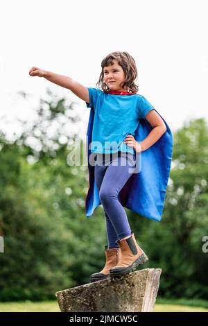 Mutiges kleines Mädchen im Superhelden-Kostüm, das geballte Fäuste hebt und brüllt, während es auf einem Steinblock steht und den Sieg im Park feiert Stockfoto