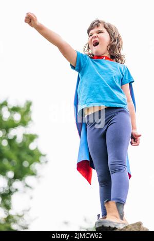 Mutiges kleines Mädchen im Superhelden-Kostüm, das geballte Fäuste hebt und brüllt, während es auf einem Steinblock steht und den Sieg im Park feiert Stockfoto