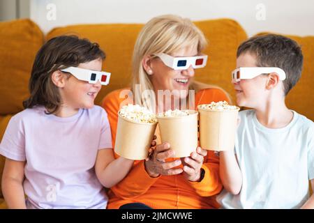 Positive Mutter und Kinder in 3D Gläsern, die leckeres Popcorn halten und sich gegenseitig angucken, während sie sich auf der Couch im Wohnzimmer einen Film ansehen Stockfoto