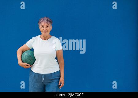 Positive ältere Frau mit kurzen Haaren in Denim-Shorts und weißem T-Shirt, das vor blauem Hintergrund mit Ball in der Hand steht und vor der Kamera lächelt Stockfoto