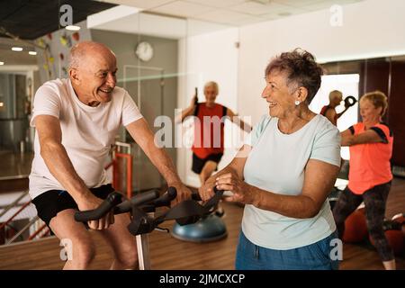 Fröhliche ältere Frau im aktiven Ohr lachend, während sie in der Nähe eines motivierten Mannes steht, der während des Trainings im modernen Fitnessstudio auf dem Radfahrgerät trainiert Stockfoto