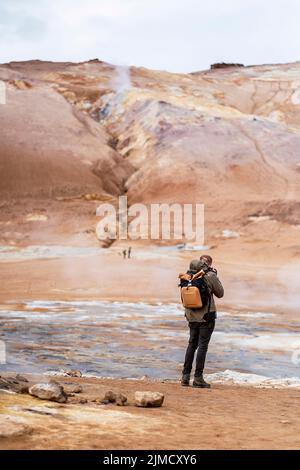 Rückansicht eines nicht erkennbaren männlichen Reisenden in zwanglosem Outfit und Rucksack, der während der Reise Geysir mit Dampf in gebirgigem vulkanischem Gelände fotografiert Stockfoto