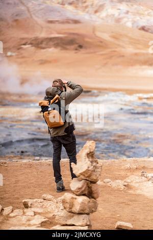 Rückansicht eines nicht erkennbaren männlichen Reisenden in zwanglosem Outfit und Rucksack, der während der Reise Geysir mit Dampf in gebirgigem vulkanischem Gelände fotografiert Stockfoto