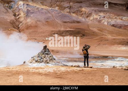 Rückansicht eines nicht erkennbaren männlichen Reisenden in zwanglosem Outfit und Rucksack, der während der Reise Geysir mit Dampf in gebirgigem vulkanischem Gelände fotografiert Stockfoto