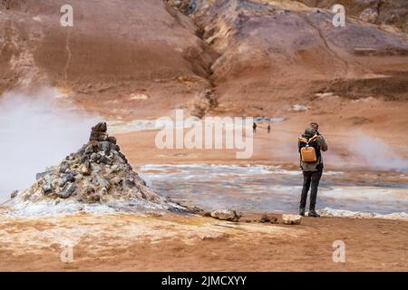 Rückansicht eines nicht erkennbaren männlichen Reisenden in zwanglosem Outfit und Rucksack, der während der Reise Geysir mit Dampf in gebirgigem vulkanischem Gelände fotografiert Stockfoto