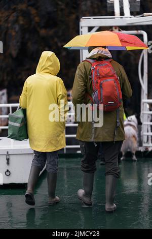 Rückansicht von nicht erkennbaren Touristen in Regenmänteln mit Regenschirm, die an regnerischen Tagen auf dem nassen Deck eines modernen Schiffes in Island spazieren gehen Stockfoto