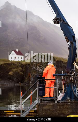 Anonymer Mann in orangefarbener Uniform, der den Kran während der Arbeit auf der Fischfarm am grauen Tag in den Bergen Islands kontrolliert Stockfoto