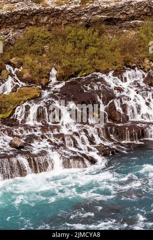 Atemberaubende Drohnenansicht des mächtigen Hraunfossar-Wasserfalls, der durch felsige Klippen in den türkisfarbenen Hvita-Fluss in Island fließt Stockfoto