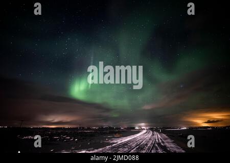 Gerade Straße durch verschneite Landschaft in Richtung Berge gegen Himmel mit grünen Polarlichtern in der Winternacht in Island Stockfoto