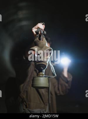 Nicht erkennbarer männlicher Entdecker in Gasmaske, der während der Apokalypse einen dunklen unterirdischen Tunnel mit Taschenlampe beleuchtet Stockfoto