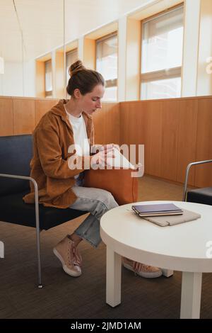 Ganzkörperschülerin in legerer Kleidung mit Haarbüschen, die Briefpapier aus der Tasche nimmt, während sie auf einem Stuhl neben dem Tisch sitzt und sich auf das Studium vorbereitet Stockfoto