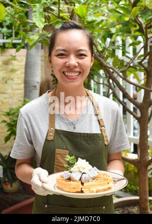 Die vietnamesische Kellnerin serviert belgische Waffeln mit Obst und Eis im Café im Freien Stockfoto