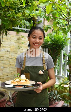Die vietnamesische Kellnerin serviert belgische Waffeln mit Obst und Eis im Café im Freien Stockfoto