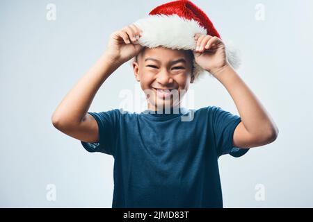 Berichterstattung für Weihnachts-Elf-Pflicht. Studioaufnahme eines niedlichen kleinen Jungen, der einen Weihnachtsmann-Hut vor einem grauen Hintergrund trägt. Stockfoto