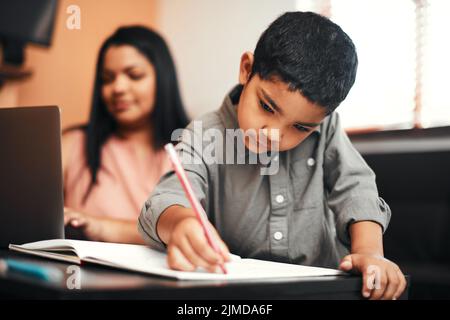 Gib ihm ein paar Buntstifte und die Hölle werde kreativ. Ein entzückender kleiner Junge färbt sich zu Hause mit seiner Mutter, die einen Laptop im Hintergrund verwendet. Stockfoto