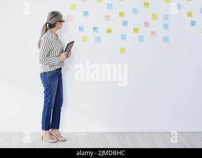 Shes arbeitete an etwas großem. Eine reife Geschäftsfrau, die ein digitales Tablet benutzte, während sie in einem Büro mit Notizen an einer Wand Brainstorming. Stockfoto