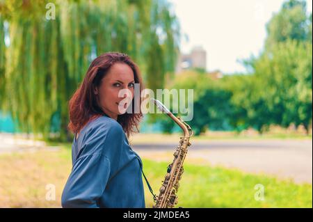 Rotes Mädchen in blauer Jacke mit einem Saxophon, das im Park lächelt Stockfoto