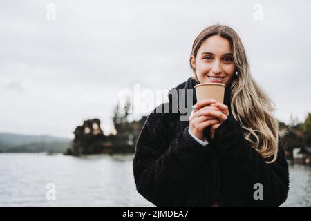 Lächelnde blonde Frau, die an einem bewölkten Nachmittag eine Einweg-Kaffeetasse vor einem schönen See hält. Modell Stockfoto