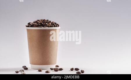 Treffen wir uns zum Kaffee. Studioaufnahme einer Papiertasse, gefüllt mit Kaffeebohnen vor grauem Hintergrund. Stockfoto