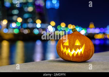 Halloween Kürbis auf dem Hintergrund der Nachtstadt. Verschwommene farbige Lichter werden im Wasser reflektiert. Nächtliches Stadtdekor mit einem Stockfoto