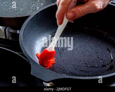 Menschliche Hand, die eine rot-heiße Pfanne mit einer roten Silikonbürste einölt. Vorbereitung zum Backen von Pfannkuchen in der heimischen Küche. Stockfoto