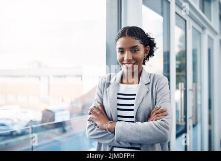 Glückliche, selbstbewusste und professionelle junge Geschäftsfrau, die ihre Arme faltete, während sie in einem modernen Büro stand. Porträt einer schwarzen Führerin, die lächelt Stockfoto